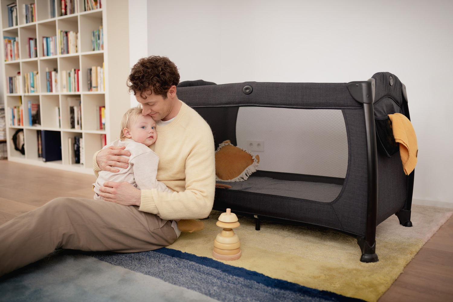 Dad cradling newborn sat next to the Hauck black travel cot in the nursery | Baby Travel Essentials | Nursery Furniture - Clair de Lune UK