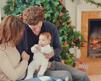 Mum, dad and baby dressed in a onsie sat next to the Christmas tree and an opne log fire | Family Christmas - Clair de Lune UK