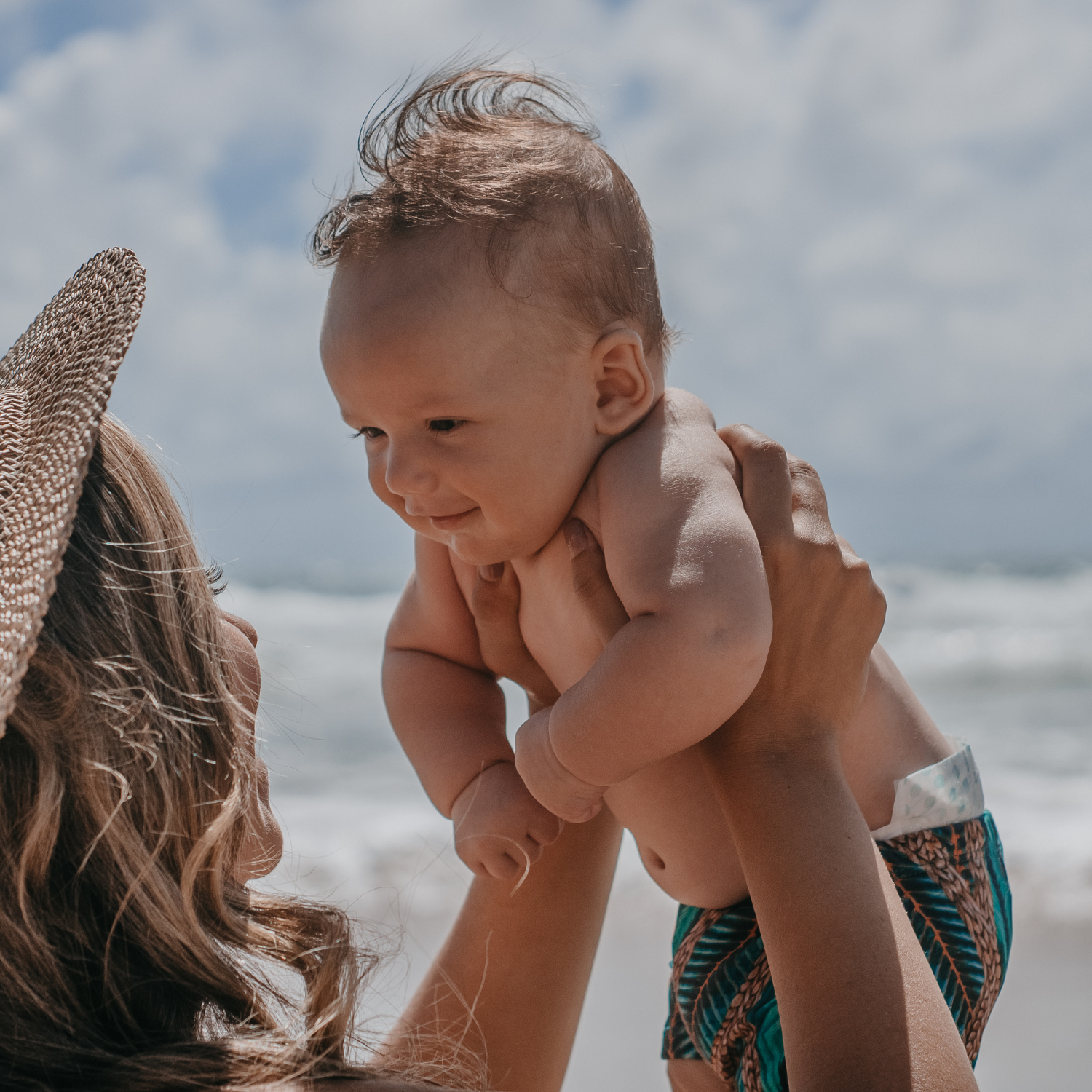 Mum wearing a straw hat holding her baby up in the air at the beach | Baby Travel Essentials - Clair de Lune UK