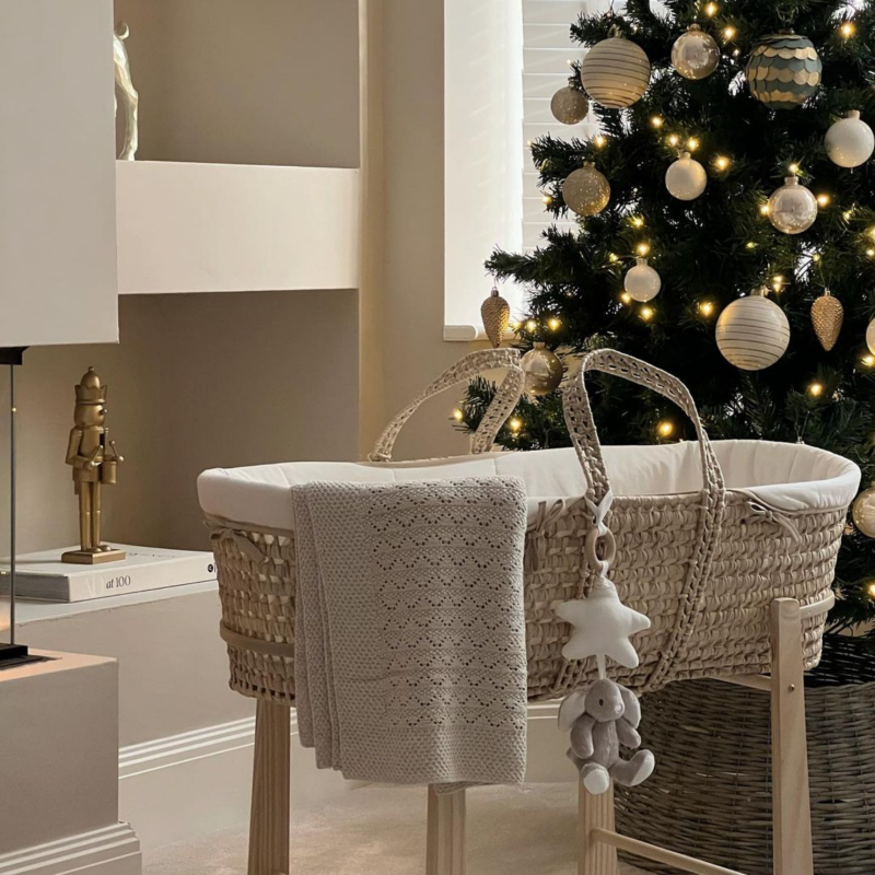 The Cream Organic Palm Moses Basket on Rocking Stand with toys attached next to a green Christmas tree with gold and beige baubles | Moses Baby Baskets, Bassinets | Baby Christmas Gifts 2024 - Clair de Lune UK