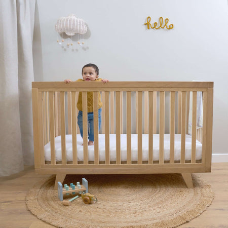 Baby standing on the cot bed mattress in the Oak Cot Bed in a Scandi Natural Nursery Room | Cots, Cot Beds, Toddler & Kid Beds | Nursery Furniture - Clair de Lune UK