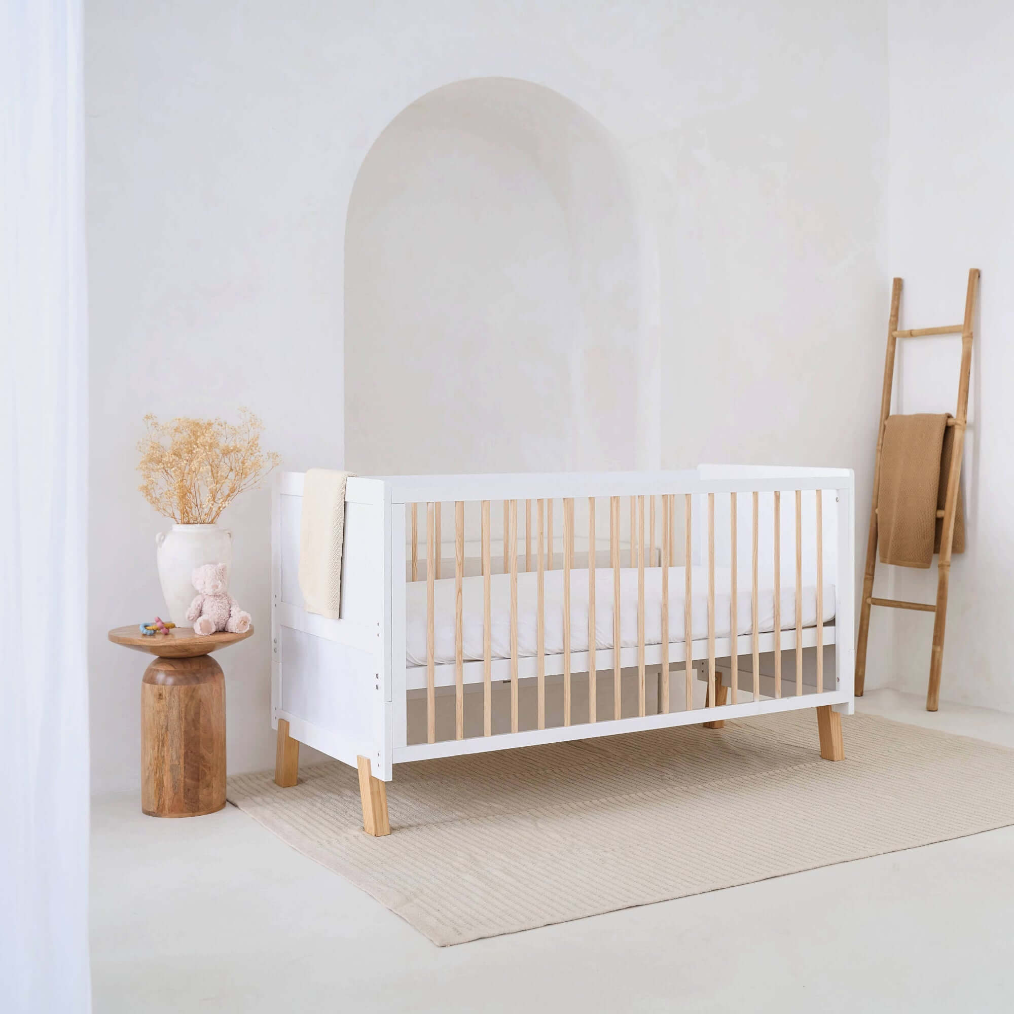White Essentials Cot Bed in a super stylish white minimalist nursery room by a wooden night stand and ladder for the organic knit blanket | Cots, Cot Beds & Toddler Beds | Nursery Furniture - Clair de Lune UK