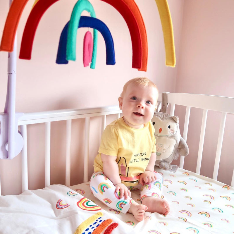 Baby playing with the Rainbow Ickle Bubba Musical Cot Mobile on an Ickle Bubba cot in a rainbow-themed nursery room | Nursery Accessories | Baby and Toddler Bedding - Clair de Lune UK