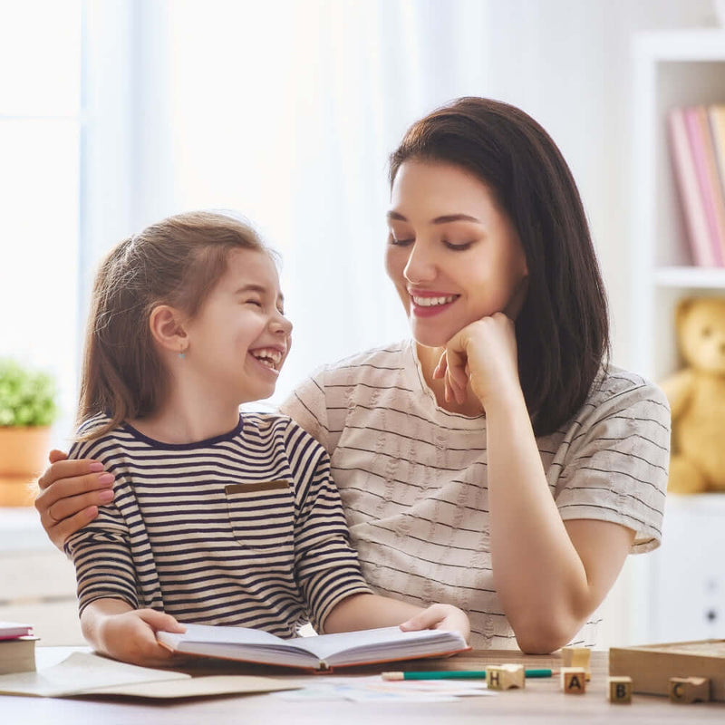 Mum and daughter with dark hair sat reading at the dining table and smiling at each other | Parenting tips and advice
