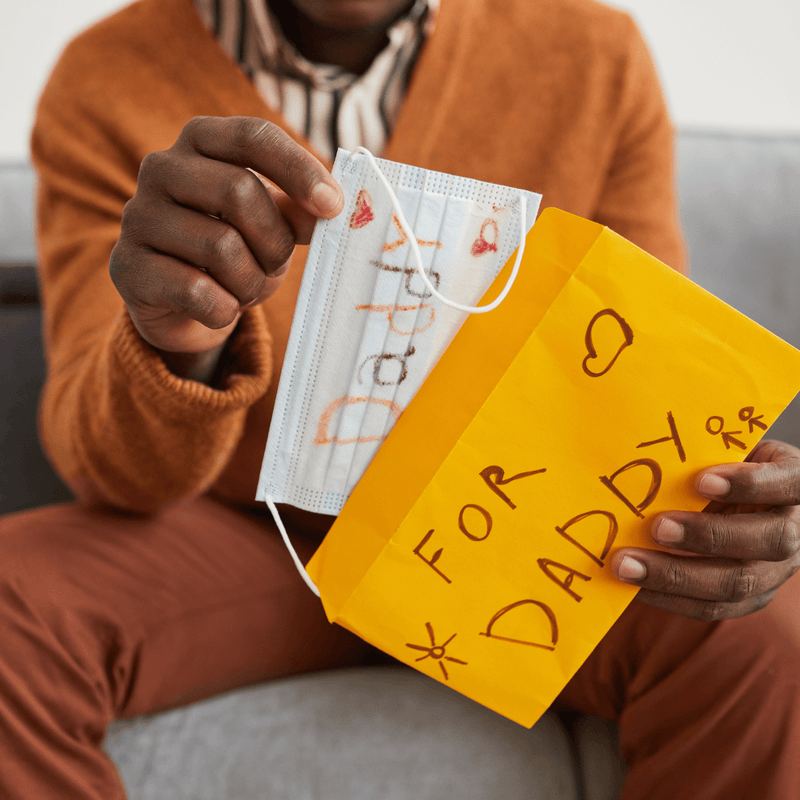 Dad wearing a mustard yellow jacket sat on a grey sofa holding his homemade Father's Day card from his child | Family Time - Clair de Lune UK