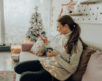 Mum sat on the sofa with a cup of tea online Christmas shopping on her phone next to the Christmas tree | Family Christmas - Clair de Lune UK