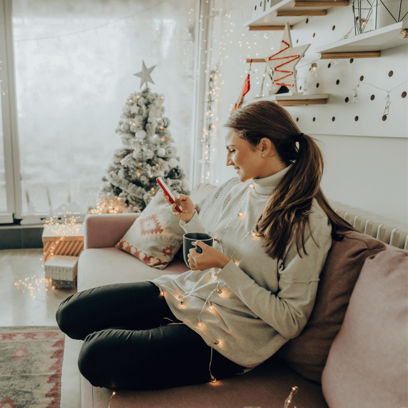 Mum sat on the sofa with a cup of tea online Christmas shopping on her phone next to the Christmas tree | Family Christmas - Clair de Lune UK