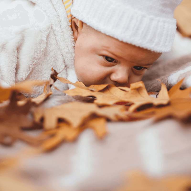 Newborn baby wearing a white knitted hat lying on their tummy on brown autumn leaves | Top 10 Autumn-Inspired Baby Names | News - Clair de Lune UK