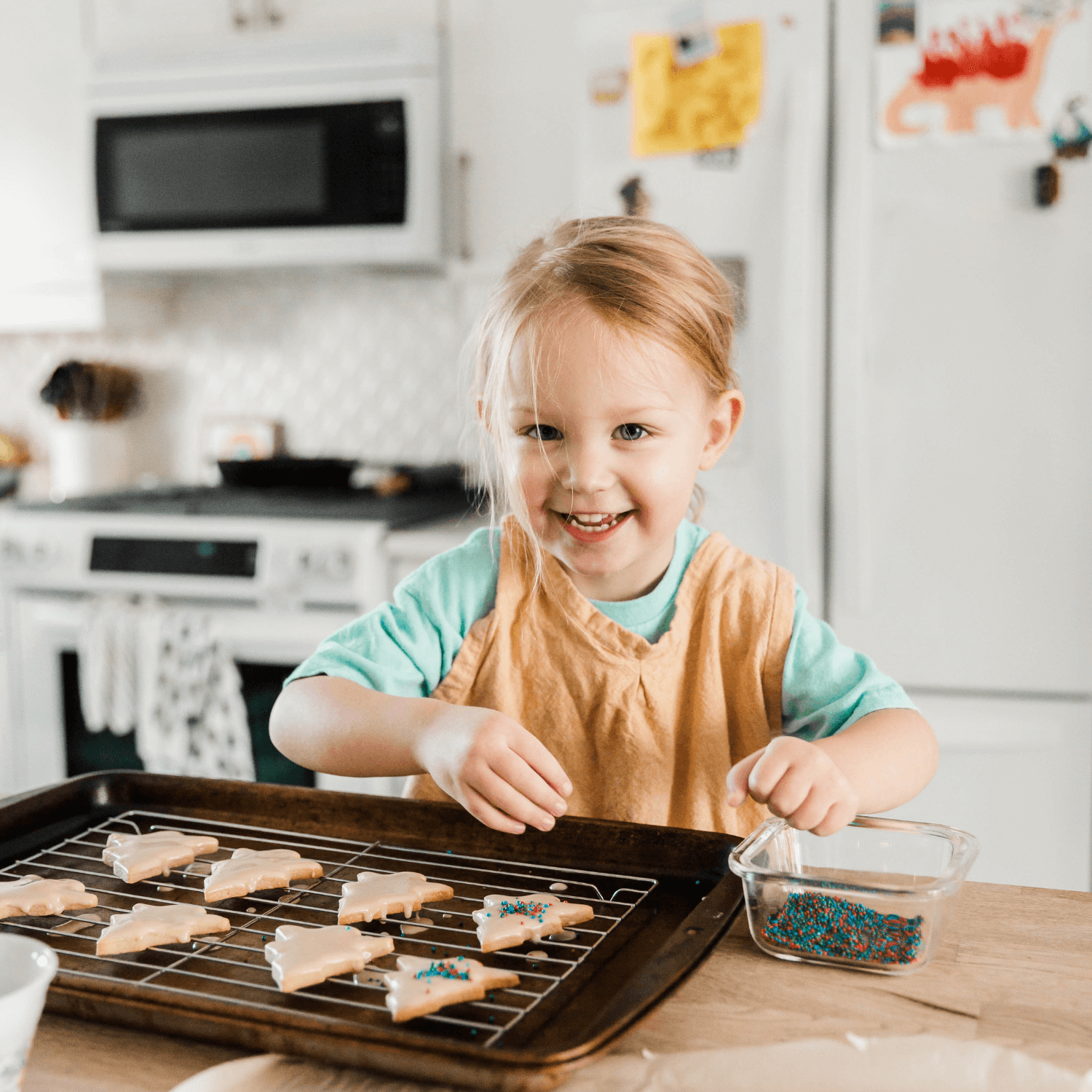 Blonde little girl stood at the kitchen island decorating Christmas tree shaped short breads with icing and sprinkles | Family Time - Clair de Lune UK