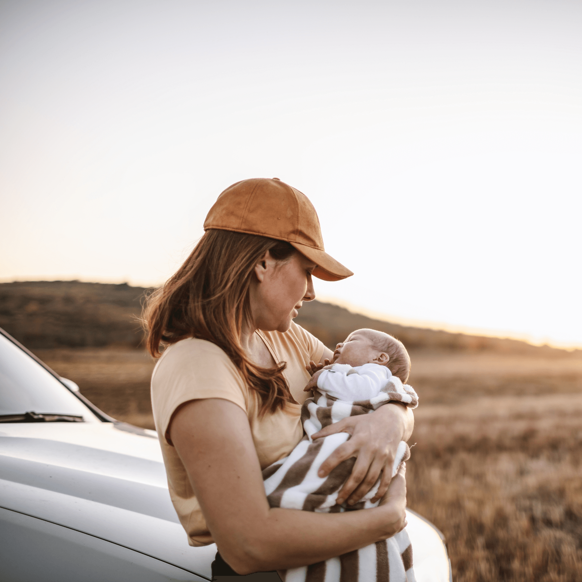 Mum wearing a brown cap stood next to a silver car holding her newborn wrapped in a cellular blanket | Travel with Baby | Family Time - Clair de Lune UK