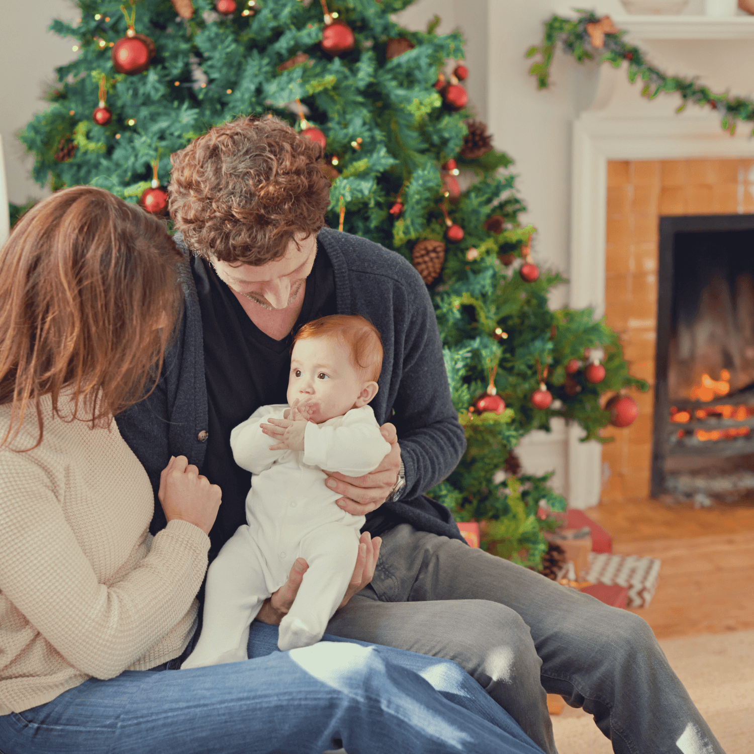 Mum, dad and baby dressed in a onsie sat next to the Christmas tree and an opne log fire | Family Christmas - Clair de Lune UK