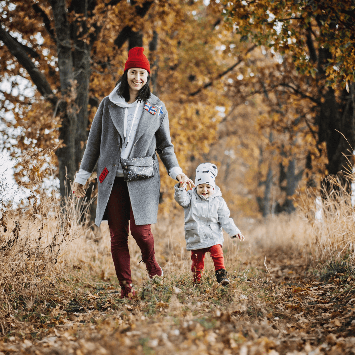 Mum and toddler girl wrapped up in coats and hats holding hands, walking down a path with autumn leaves and trees | Autumn Baby Activities: Top 5 Outdoor Fun Ideas - Clair de Lune UK