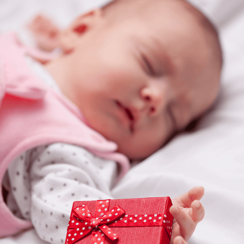 Newborn baby lying asleep on the bed next to a Christmas gift wrapped in red | Family Christmas - Clair de Lune UK