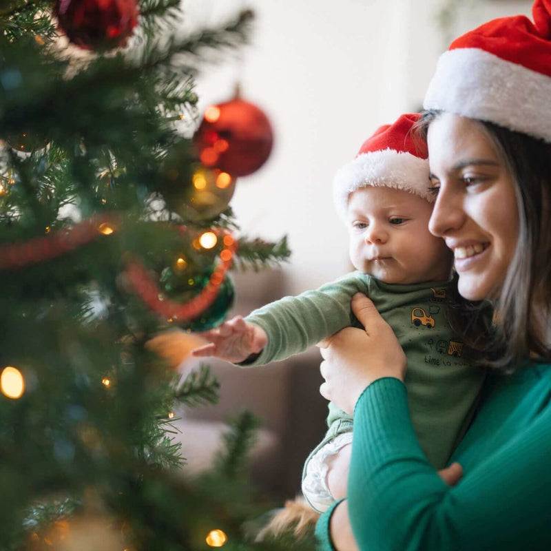 Mum holding newborn baby and showing them the christmas tree while wearing santa hats | Christmas Eve Traditions - Clair de Lune UK