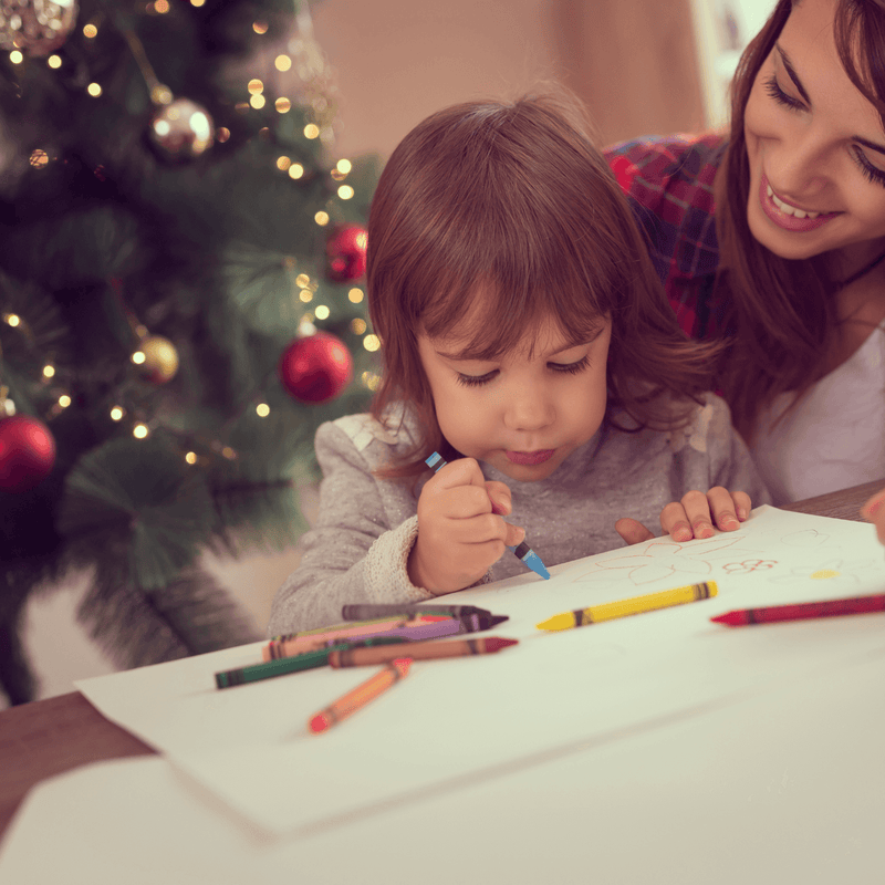 Dark haired toddler and mum sat at the table next to the Christmas tree drawing and colouring Christmas cards | Family Christmas - Clair de Lune UK