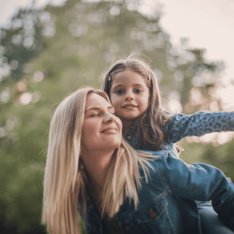 Happy mum giving daughter a piggy back in the sunny park  - Clair de Lune UK