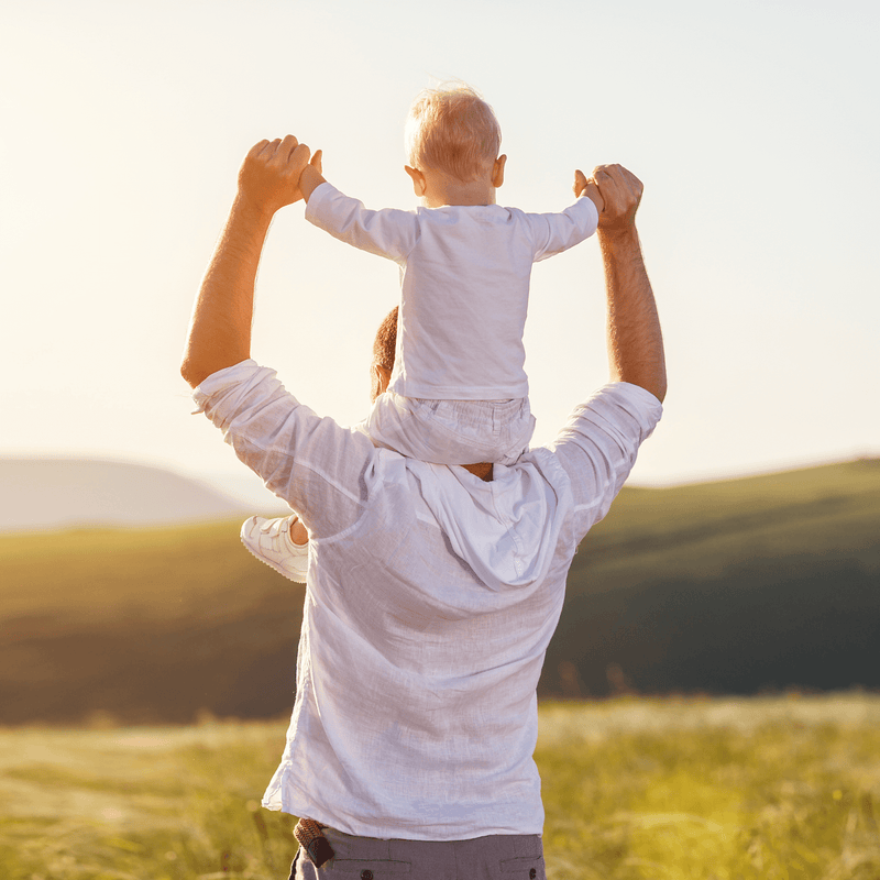 Dad wearing shirt and jeans with his blonde haired son wearing a white shirt sat on his shoulders holding hands looking out over a park | Faminly Time | Father's Day - Clair de Lune UK