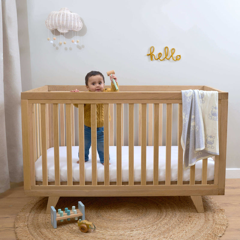Clair de Lune Oak Cot Bed | Toddler boy standing in the Natural Oak Cot Bed in baby room | Nursery Furniture - Clair de Lune UK