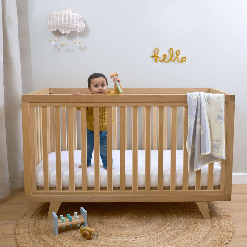 Toddler boy standing in the Natural Oak Cot Bed in baby room | Nursery Furniture - Clair de Lune UK