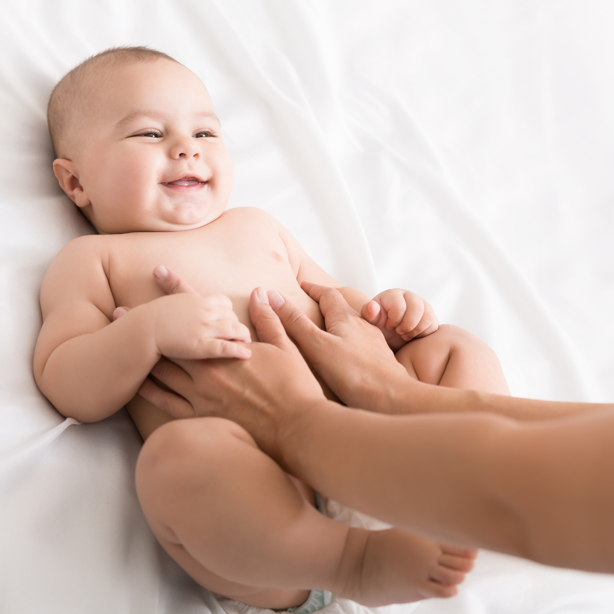 Baby lying on a white sheet with mums hands on their tummy | Parenting tips and advice | Colic Awareness - Clair de Lune UK
