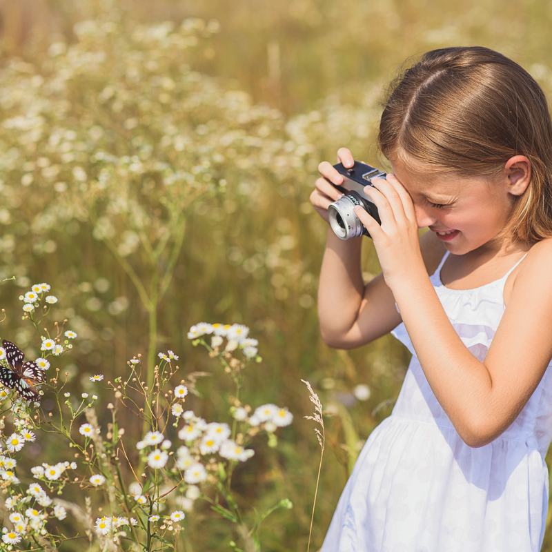 Girl wearing a white dress in a field taking a close up photo of a butterfly on a white flower | Kids Activities | Family Time - Clair de Lune UK