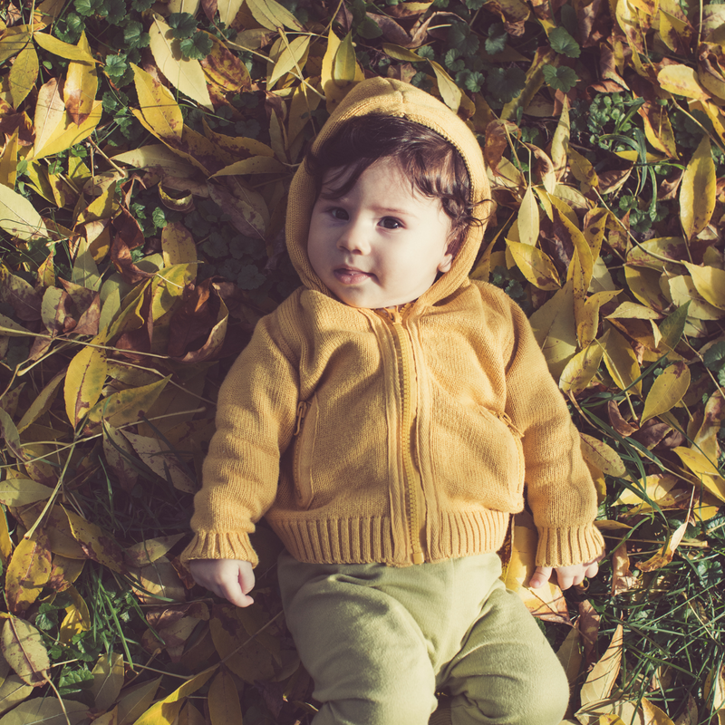 Dark haired baby boy wearing a mustard coat lying on a pile of Autumn leaves smiling - Clair de Lune UK