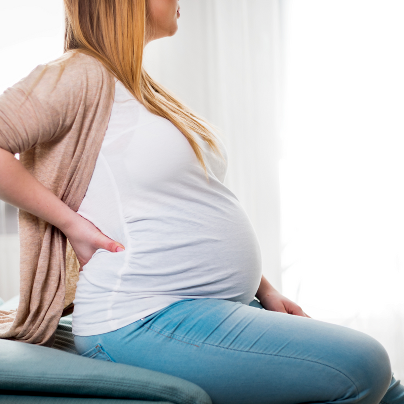 Heavily pregnant mum wearing white t-shirt and jeans sat on a chair holding her back | Pregnancy - Clair de Lune UK