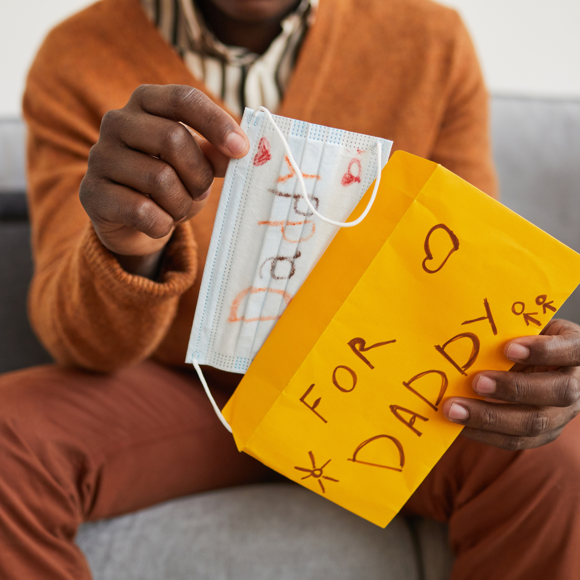 Dad wearing a mustard yellow jacket sat on a grey sofa holding his homemade Father's Day card from his child | Family Time - Clair de Lune UK