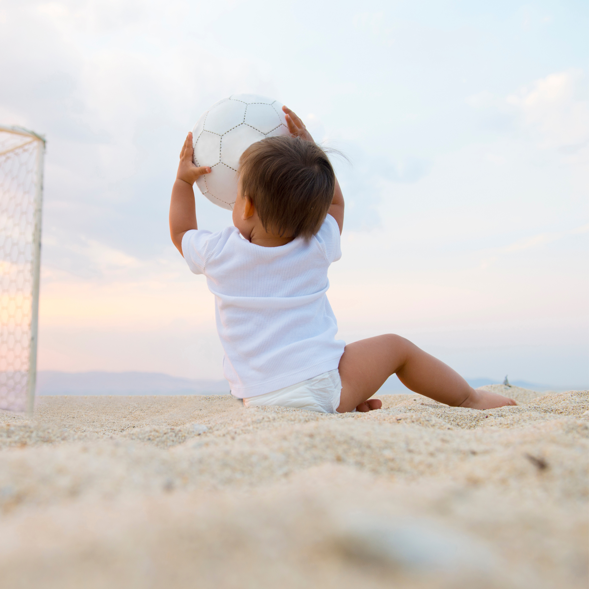 Toddler wearing a nappy and a white t-shirt sat on the beach holding a football | Summer Safety - Clair de Lune UK