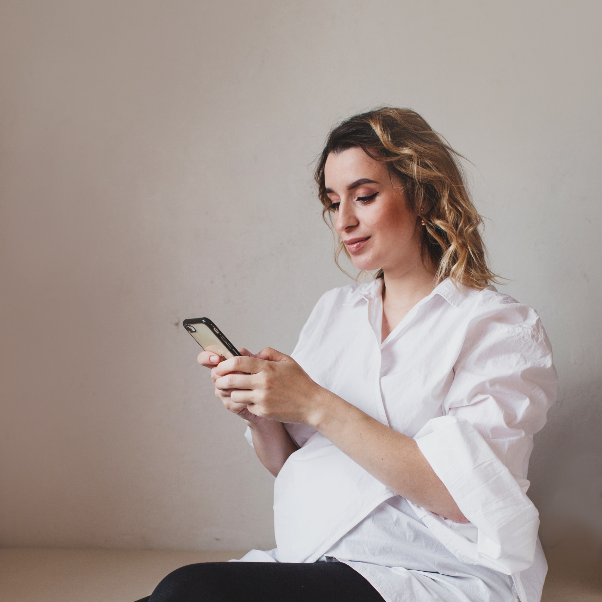 Pregnant mum wearing a white shirt against a grey back drop scrolling on her phone | Pregnancy Tips - Clair de Lune UK