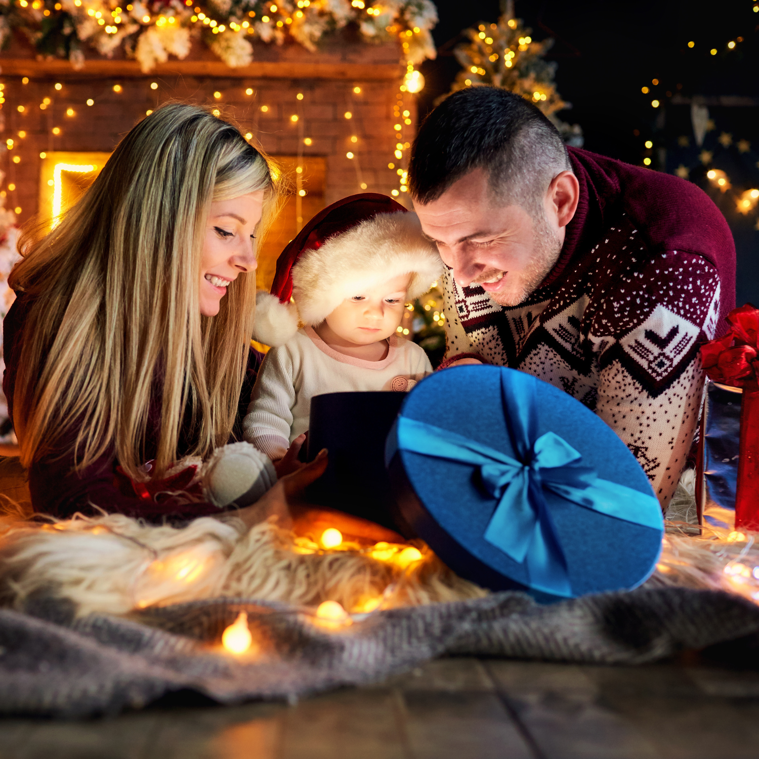 Mum, dad and baby wearing a santa hat looking inside a gift box surrounded by twinkling fairy lights | Family Christmas - Clair de Lune UK