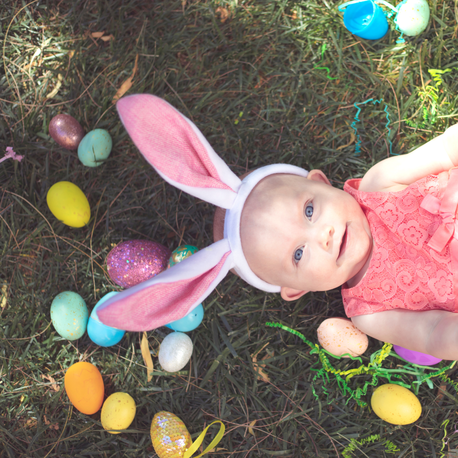 Toddler girl wearing bunny ears lying on the grass surrounded by plastic, fillable Easter eggs - Clair de Lune UK