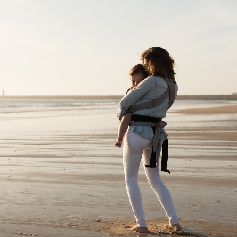 Mum wearing jeans and a jumper holding her toddler in a harness at the beach | Travel with Baby | Family Time - Clair de Lune UK