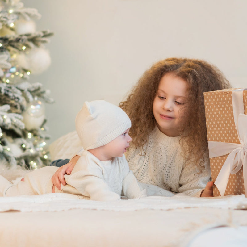 Baby and toddler lying by the Christmas tree on a white fur rug opening a Christmas present | Toddler and Baby Christmas Gifts | Baby & Toddler Gifts & Toys - Clair de Lune UK