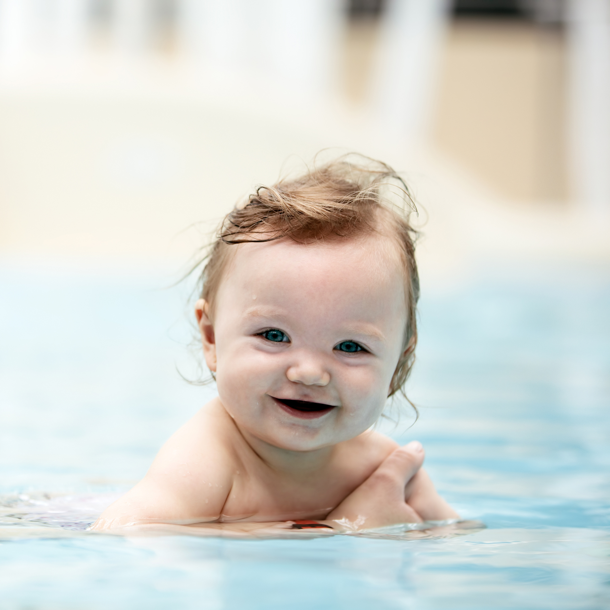 Smiling baby with long brown hair being held in the swimming pool | Water Safety | Family Time - Clair de Lune UK