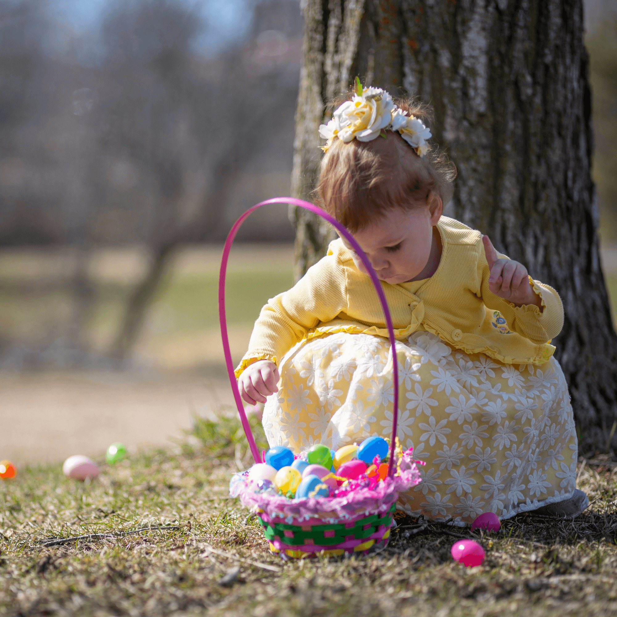 Toddler wearing a pretty flower dress, yellow cardigan and daisy headband squatting down adding plastic eggs to her Easter basket | Easter Kids Activities | Family Time - Clair de Lune UK