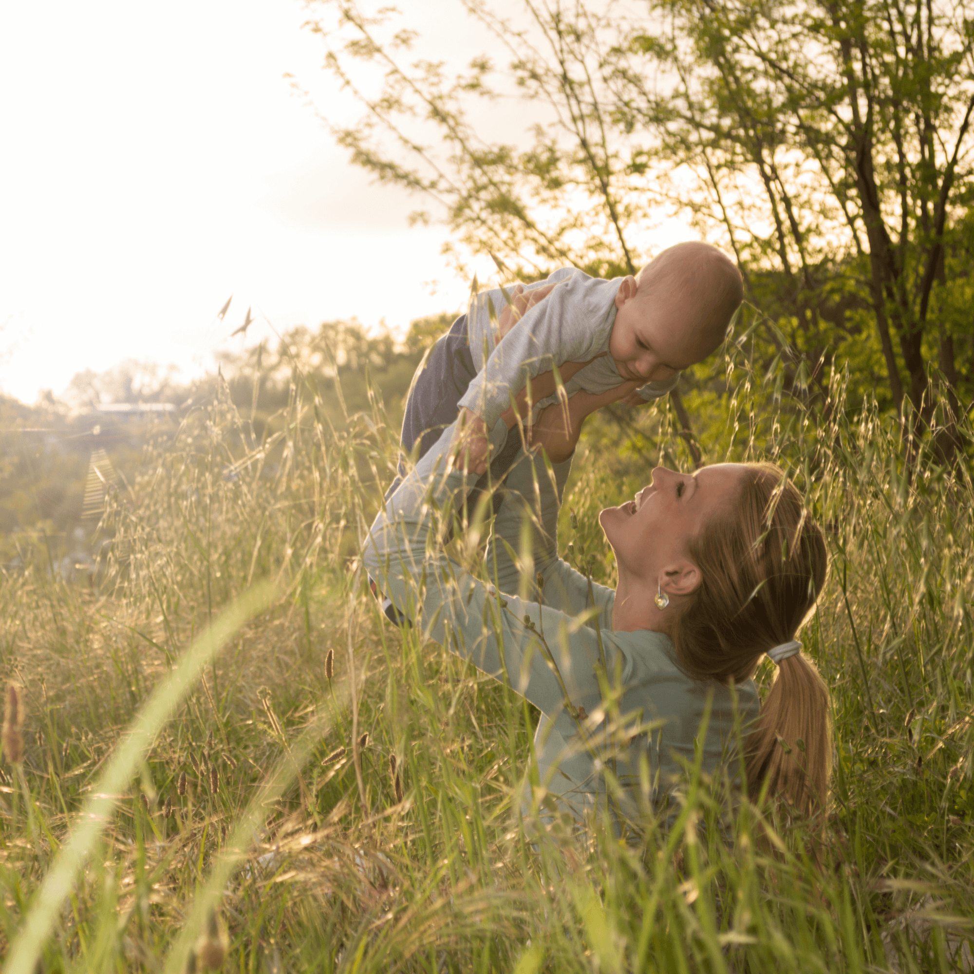 Mum with hair in a ponytail sitting in the long grass holding her baby son above her head and smiling | Baby Name Inspiration - Clair de Lune UK