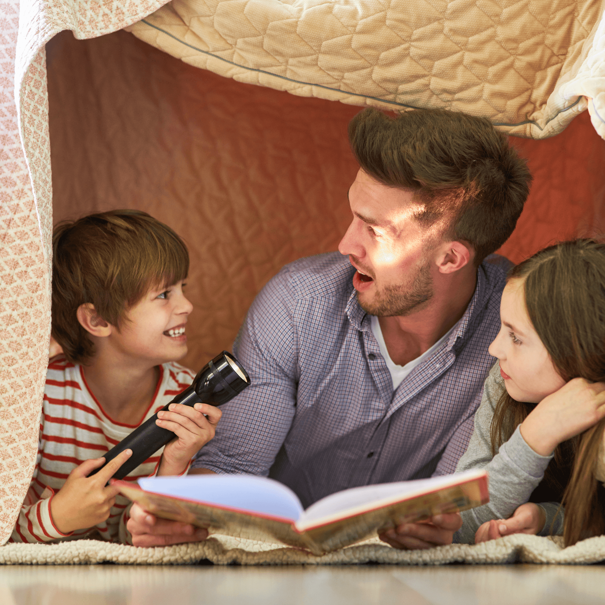 Dad and his two children lying on their stomachs in a blanket fort reading a story | Family Time - Clair de Lune UK