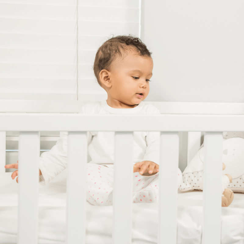 Dark haired baby wearing a white baby grow in the Essentials Cot Bed | Nursery Furniture | Baby Essentials - Clair de Lune UK