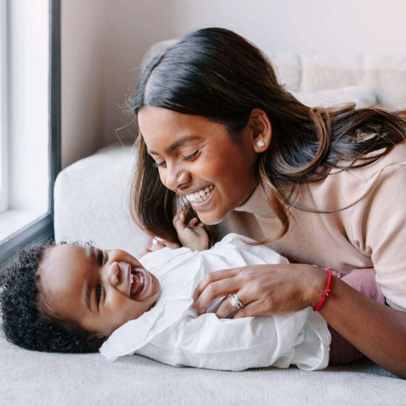 Mum and toddler laughing while tickling each other on the bed by a window | The First Mother's Day | Parenting Advice - Clair de Lune UK