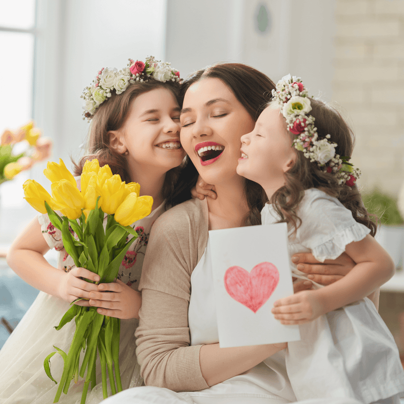 Mum smiling with her two daughters both wearing flower garlands gifting their mum a bunch of daffodils and a homemade card with a heart on | Family Time | Mother's Day - Clair de Lune UK