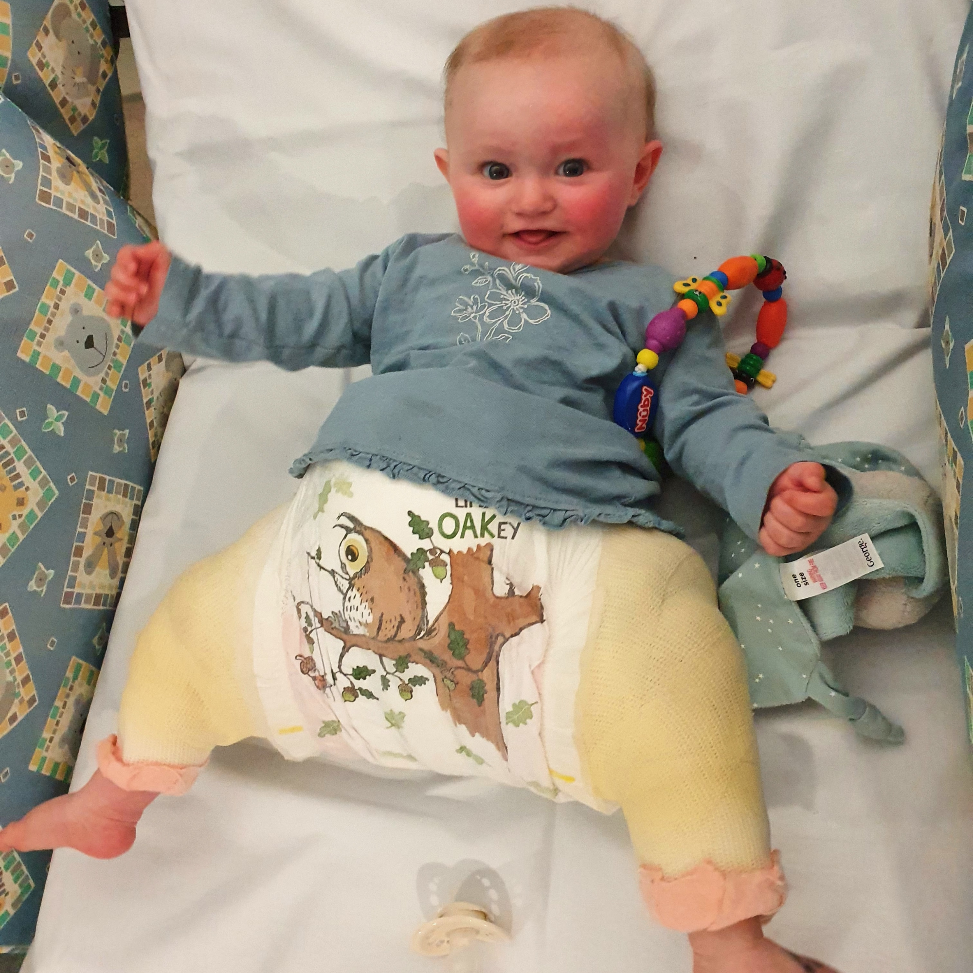 Beautiful 4 month old baby lying in a hospital bed wearing a hip spica cast playing with her toys | Hip Dysplasia - Clair de Lune UK