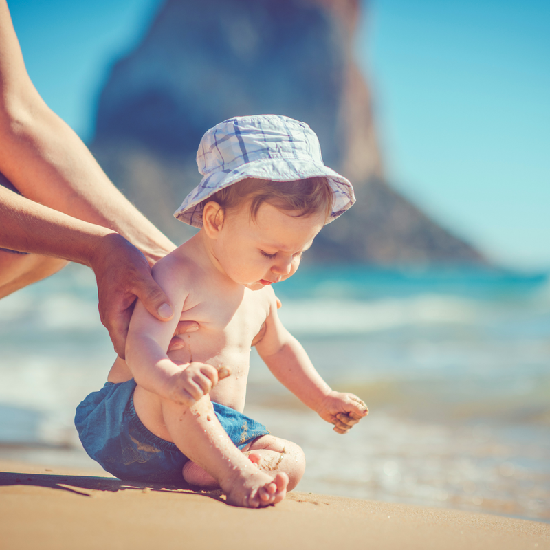 Baby boy wearing a bucket hat and swim shorts playing in the sand at the beach | Baby travel essentials - Clair de Lune UK