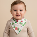 A smiling baby wearing the GrowGrows Alpine Adventures Bib with a green pine tree and red cabin pattern, paired with a beige ribbed long-sleeve top, set against a solid beige background. | Bamboo Babywear - Clair de Lune UK