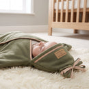 A baby swaddled in the Sage Star Fleece Baby Wrap Blanket, lying on a white shag rug in a softly lit nursery next to a wooden crib. | Cosy Baby Blankets - Clair de Lune UK