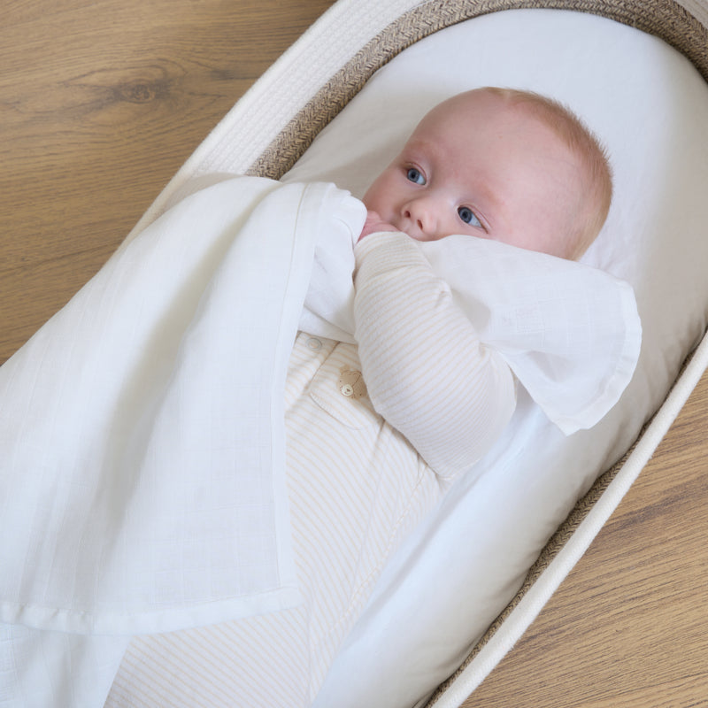 Newborn lying in a bassinet holding a 70 x 70 cm white muslin cloth and smiling | Baby Feeding & Weaning - Clair de Lune UK