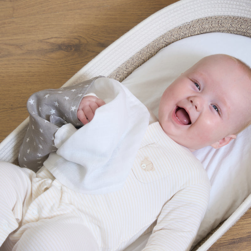 Newborn baby lying in the Moses basket smiling in a white onesie holding a grey muslin square | Feeding Essentials | Feeding & Weaning | Toddler Essentials - Clair de Lune UK