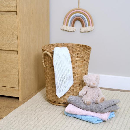 White Cotton Cellular Blanket draped over a hyacinth toy basket in the nursery next to a stack of cellular pram blankets with a teddy bear | Cosy Baby Blankets | Swaddles | Nursery Bedding - Clair de Lune UK