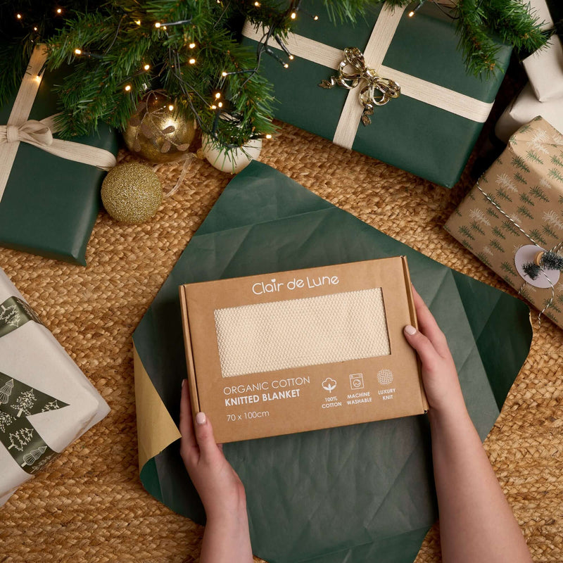 Person holding a Clair de Lune organic cotton knitted blanket box under a decorated Christmas tree | Soft Baby Blankets | Organic Nursery Essentials | Baby's First Christmas - Clair de Lune UK
