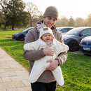 A smiling dad outdoors in a park holding a baby wrapped in the cram Star Fleece Baby Wrap Blanket. The dad is wearing a brown jacket and a beige beanie, and the background shows a grassy lawn, a stone path, and parked cars. | Cosy Baby Blankets - Clair de Lune UK